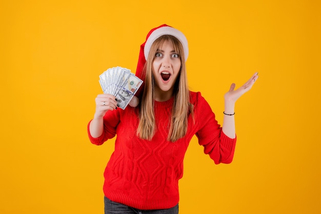 Ragazza sorpresa che tiene in mano cento banconote in dollari che porta il cappello di Santa