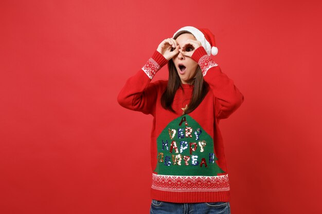 Ragazza sorpresa di Santa che tiene la bocca aperta, tenendosi per mano vicino agli occhi, imitando occhiali o binocoli isolati su sfondo rosso. Felice Anno Nuovo 2019 celebrazione festa concetto. Mock up copia spazio.