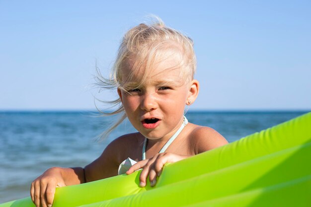 Ragazza sorridente allegra del bambino che tiene materasso gonfiabile sulla spiaggia.