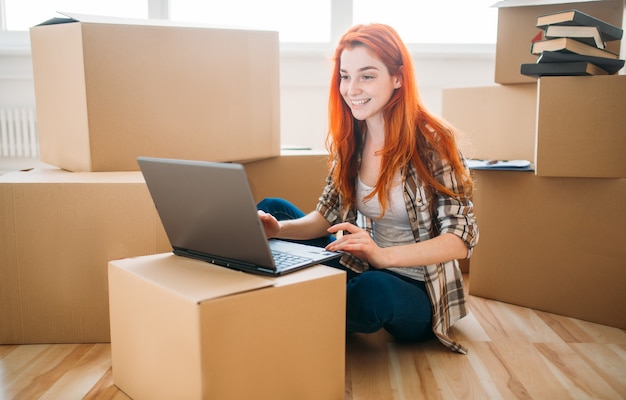 Ragazza sorridente che utilizza computer portatile tra scatole di cartone, trasferirsi in una nuova casa, inaugurazione della casa