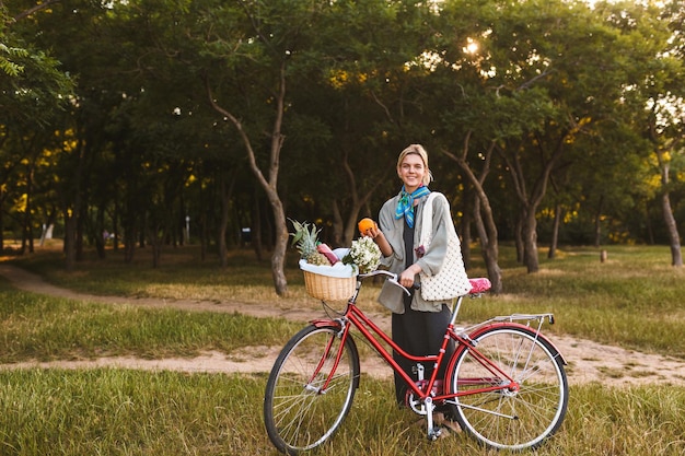 Ragazza sorridente con bicicletta rossa e cesto pieno di fiori di campo e frutta che guarda felicemente in macchina fotografica tenendo l'arancia in mano nel parco