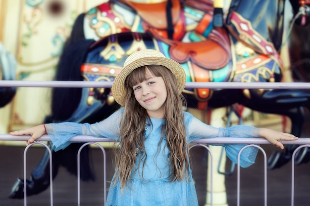 ragazza sorridente con cappello di paglia vicino alla giostra alla fiera Ritratto ragazza carina sul parco divertimenti