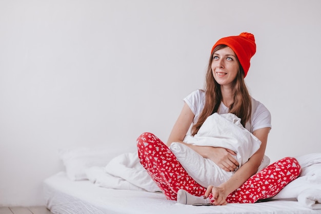 Ragazza sorridente con cappello rosso e pigiama natalizio seduta a letto con un cuscino bianco