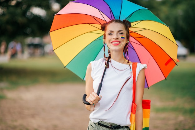 Ragazza sorridente con le trecce, la bandiera del lgbt sul suo fronte, la borsa variopinta e la posa dell'ombrello all'aperto con il fronte sorridente felice.