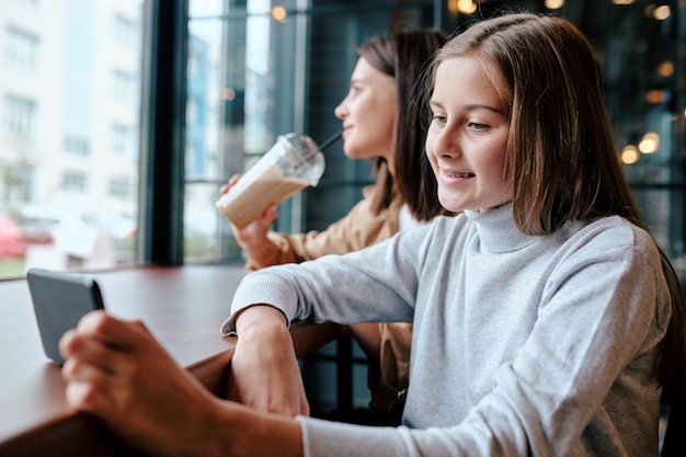 Ragazza sorridente con lo smartphone seduti a tavola nella caffetteria e guardare video con sua madre che mangia cocktail