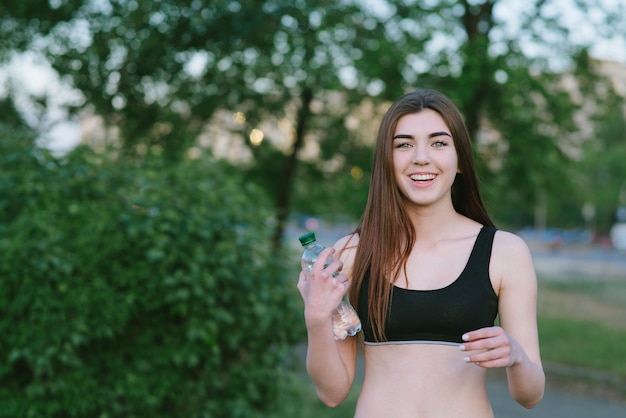 Ragazza sorridente con una figura atletica con una bottiglia di acqua a disposizione. Atleta femminile che esegue una maglietta nel parco.