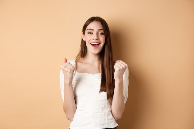 Ragazza sorridente felice che celebra la vittoria, facendo pompare il pugno e dire di sì, raggiungere l'obiettivo, trionfare o motivarsi, in piedi su sfondo beige.