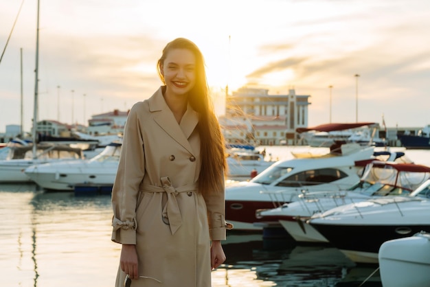 Ragazza sorridente felice con capelli lunghi in cappotto beige alla moda che posa in porto marittimo al tramonto