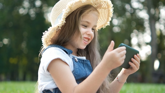 Ragazza sorridente felice del bambino che osserva nel telefono cellulare all'aperto in estate.