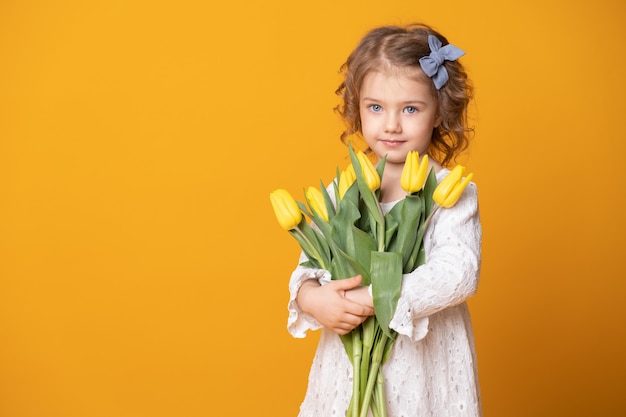 Ragazza sorridente in abito bianco su sfondo giallo. Allegro bambino felice con bouquet di fiori di tulipani.