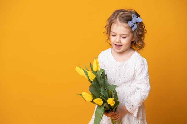 Ragazza sorridente in abito bianco su sfondo giallo. Allegro bambino felice con bouquet di fiori di tulipani.