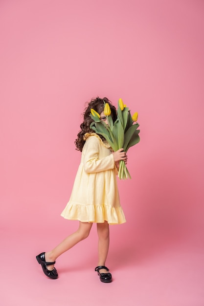 Ragazza sorridente in abito giallo su sfondo rosa studio. Allegro bambino felice con bouquet di fiori di tulipani.