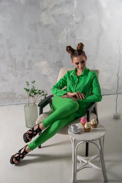 Ragazza sorridente in camicia verde e pantaloni si siede elegantemente sulla sedia in studio Modello con pose di trucco serale