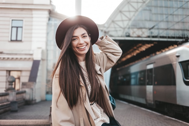 Ragazza sorridente in cappello alla partenza aspettante del treno della stazione ferroviaria