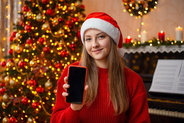 Ragazza sorridente in cappello di Babbo Natale e maglione rosso che mostra lo schermo del telefono alla fotocamera Ritratto di una giovane ragazza con il telefono