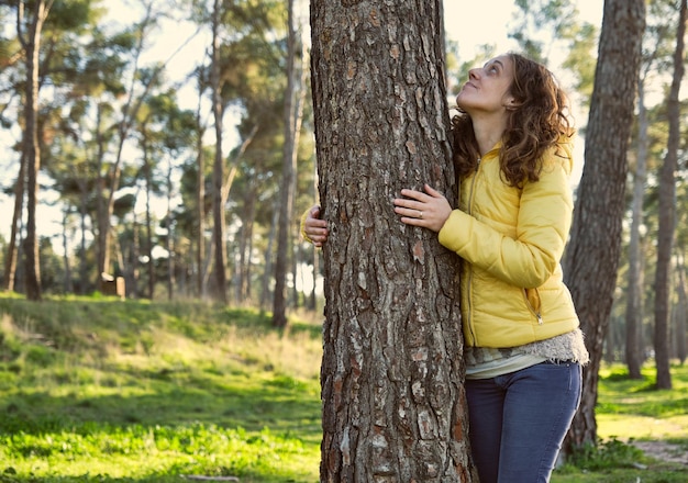 Ragazza sorridente in giacca gialla e pantaloni blu che guarda in alto e abbraccia un albero in una foresta di pini al tramonto