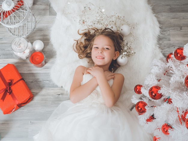 Ragazza sorridente in vestito operato bianco che si trova sul tappeto soffice tra le decorazioni di Natale