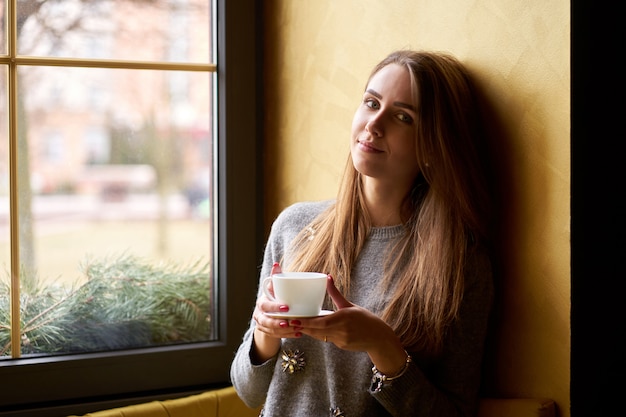 Ragazza sorridente sveglia con ubicazione dei capelli lunghi vicino alla finestra e bere caffè o tè nella caffetteria.