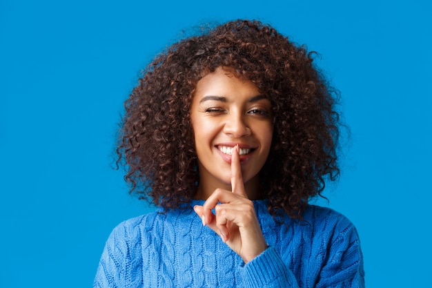 Ragazza spensierata felice afro-americana hipster, con elegante taglio di capelli afro, strizzatina d'occhio fredda e impertinente, sorridente fa zitto gesto, premi il dito indice sulle labbra mantieni la promessa di non dire segreto, prepara la sorpresa