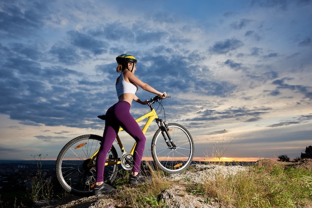 Ragazza sportiva che sta bici vicina e che posa sulla collina. Donna snella e bella. Incredibile sfondo del cielo nuvoloso. Attività sportive all'aria aperta.