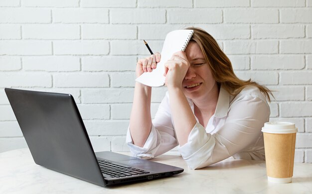 Ragazza stanca che si siede allo scrittorio di lavoro a casa che studia al computer portatile con il caffè