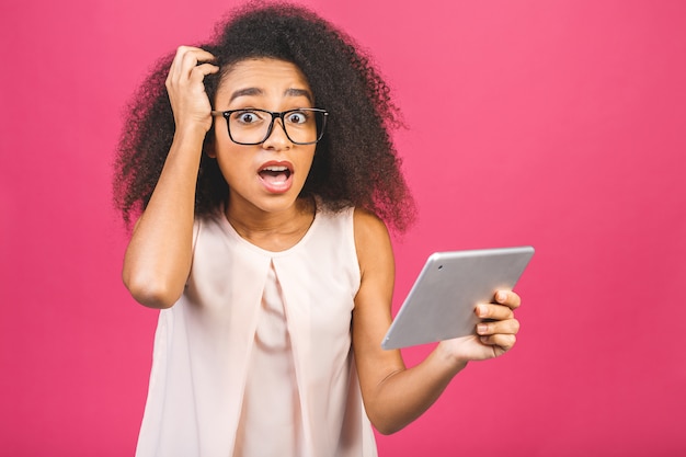 Ragazza studentessa americana scioccata con capelli africani ricci che tiene compressa digitale sopra il rosa con lo spazio della copia per testo, logo o pubblicità.