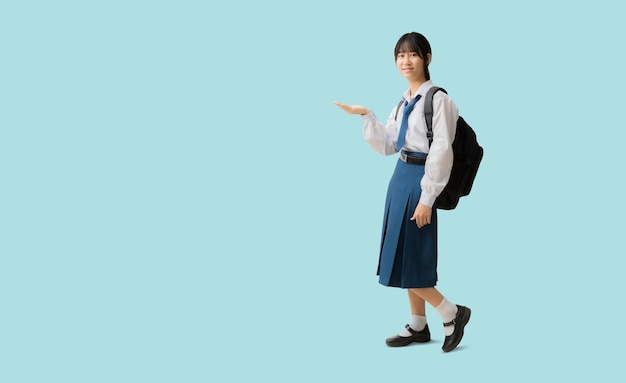 Ragazza studentessa asiatica a corpo pieno che indossa uno zaino da tenere in uniforme con la mano che tiene il palmo aperto su uno spazio vuoto isolato su sfondo blu con tracciati di ritaglio per il lavoro di progettazione vuoto spazio libero