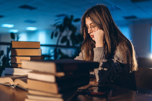 Ragazza studentessa stanca la sera seduta appoggiata al braccio in biblioteca alla scrivania con i libri. Insegnamento e preparazione agli esami.