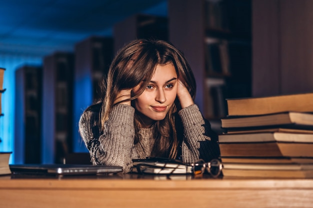 Ragazza studentessa stanca la sera seduta appoggiata al braccio in biblioteca alla scrivania con i libri. Insegnamento e preparazione agli esami