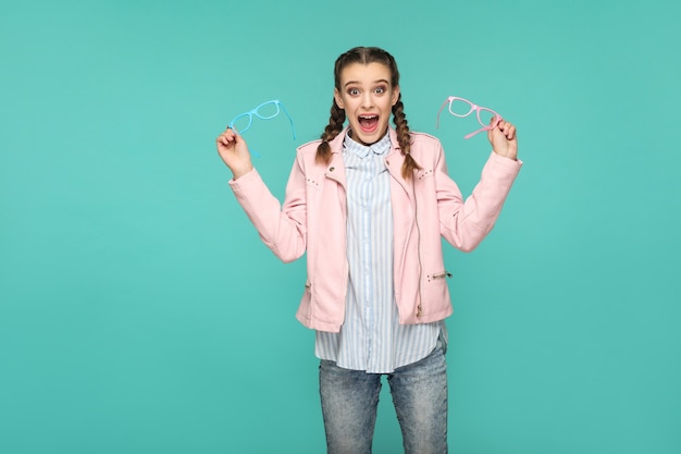 Ragazza stupita in stile casual o hipster, acconciatura a codino, in piedi, con in mano occhiali blu e rosa e guardando la telecamera con faccia sorpresa, ripresa in studio al coperto, isolata su sfondo blu o verde