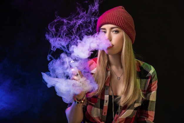 Ragazza svapata. Giovane donna hipster vape e-cig in studio su sfondo nero. Stile hip-hop. Avvicinamento.