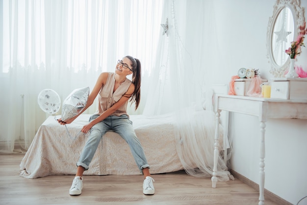 Ragazza sveglia del primo piano del brunette nel, che sorride ampiamente e che gioca con gli aerostati trasparenti e d'argento. Indossa occhiali e capelli attorcigliati.