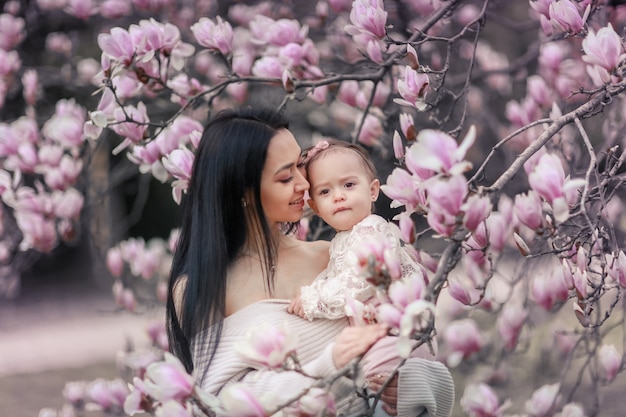 Ragazza sveglia di 6 mesi del bambino in attrezzatura rosa con i grandi occhi azzurri con la giovane bella madre alla primavera, albero di fioritura rosa ai precedenti