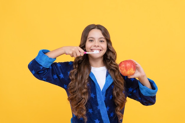 Ragazza teenager allegra con capelli ricci in pigiama comodo che lava i denti e tiene la mela, odontoiatria.