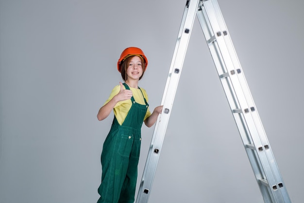 Ragazza teenager allegra in uniforme da lavoro e casco sulla scala mostra pollice in alto edificio