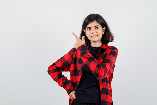 Ragazza teenager che indica all'angolo in alto a sinistra in t-shirt, camicia a scacchi e guardando gioiosa, vista frontale.