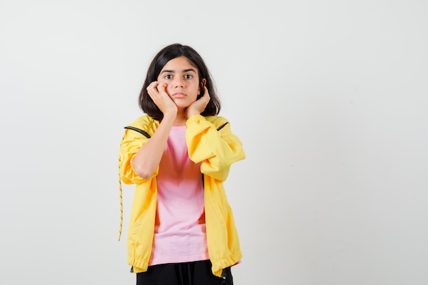 Ragazza teenager che si appoggia il mento sulle mani in tuta gialla, t-shirt e sembra sorpresa, vista frontale.