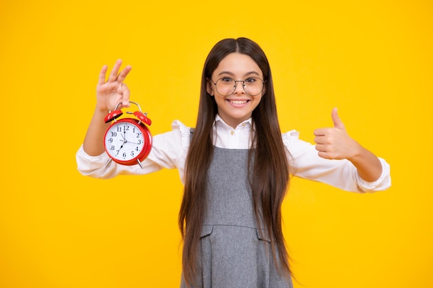 Ragazza teenager che tiene l'orologio su sfondo giallo Programma giornaliero del mattino presto Il tempo di controllo del bambino non è in ritardo Bambino puntuale con allarme Tempo di scadenza Faccia felice