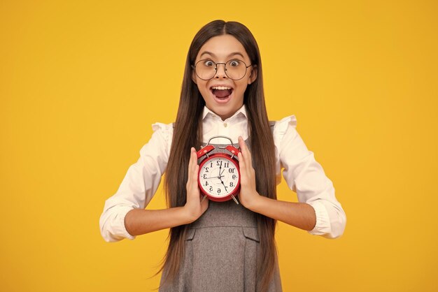 Ragazza teenager che tiene l'orologio su sfondo giallo Programma giornaliero del mattino presto Il tempo di controllo del bambino non è in ritardo Bambino puntuale con allarme Tempo di scadenza Ragazza adolescente sorpresa