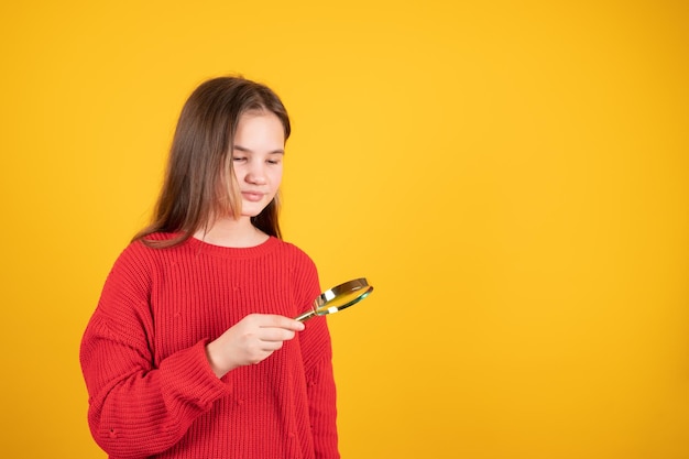 Ragazza teenager che tiene la lente d'ingrandimento e guarda qualcosa Giovane studentessa in maglione rosso su sfondo giallo