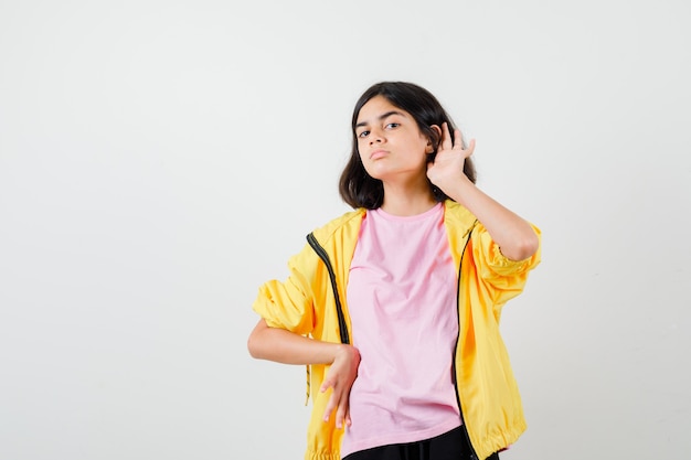 Ragazza teenager che tiene la mano dietro l'orecchio, che tiene la mano sulla vita in maglietta, giacca e sembra concentrata. vista frontale.