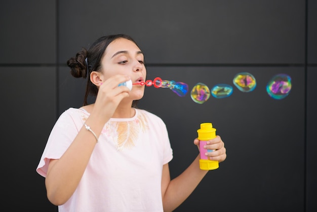 Ragazza teenager con i panini per capelli che soffiano bolle di sapone colorate mentre si gode l'estate contro il muro grigio