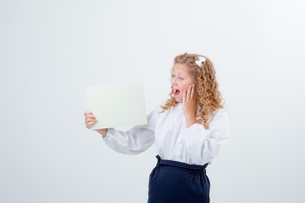 Ragazza teenager della scolaretta in uniforme scolastica che tiene un foglio di carta bianco sorpresa su una priorità bassa bianca