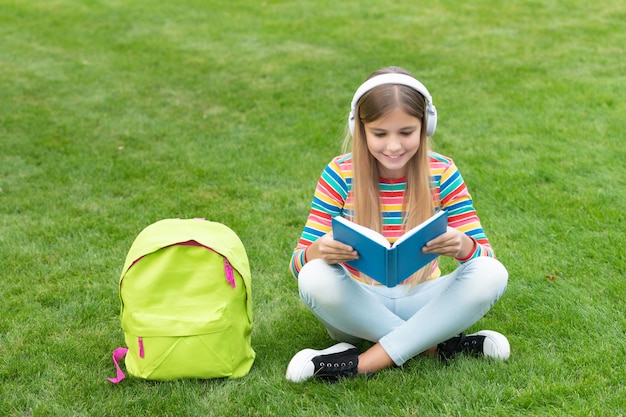 Ragazza teenager felice che legge e ascolta l'audiolibro nelle cuffie che si siede sull'erba all'aperto della scuola