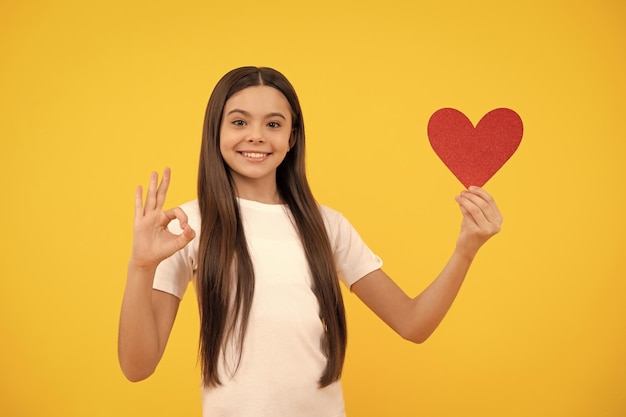 Ragazza teenager felice che tiene il cuore di biglietti di S. Valentino che mostra il gesto giusto sull'amore giallo del fondo