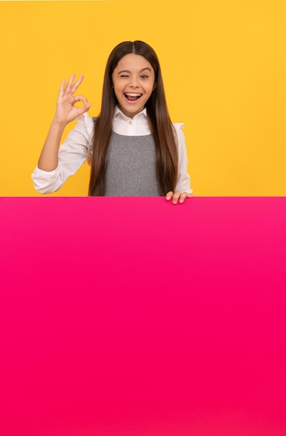 Ragazza teenager felice dietro il foglio di carta rosa in bianco per il gesto giusto dello spazio della copia, annuncio.
