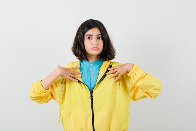 Ragazza teenager in giacca gialla che indica se stessa e sembra perplessa, vista frontale.