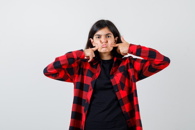 Ragazza teenager in t-shirt, camicia a scacchi premendo i pugni sulle guance e guardando divertita, vista frontale.