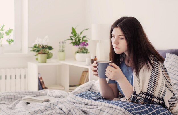 Ragazza teenager malata con una tazza di tè sul letto nella stanza bianca