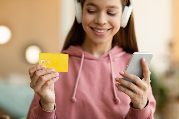 Ragazza teenager positiva in cuffia che utilizza smartphone e carta di credito per lo shopping online a casa